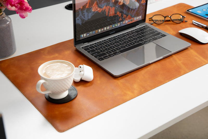 Laptop on a brown leather desk pad with a cup of coffee and earbuds on a white desk.