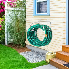 A green coiled garden hose hanging on a white metal wall-mounted hose reel attached to a beige house exterior, with grass, gravel, flowers, and wooden steps visible.
