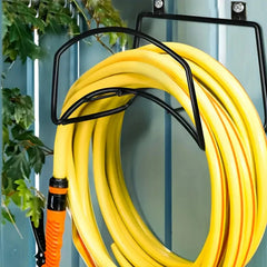 A coiled yellow garden hose hanging from a black metal wall-mounted hose rack, with an orange and black hose connector visible at the bottom left against a blue fence background.