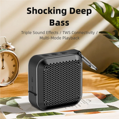 A small black portable Bluetooth speaker with a metal carabiner clip, shown on a tabletop with printed papers, a clock, and a plant in a styled indoor setting. Large text above reads 'Shocking Deep Bass' with additional feature copy.