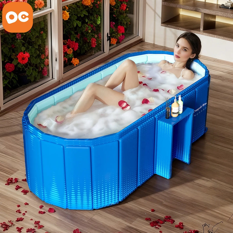 A woman relaxes in a blue foldable bathtub filled with bubbly bathwater and scattered rose petals, placed on a wooden floor near large windows with flowers outside. An orange OG logo appears in the top-left corner.
