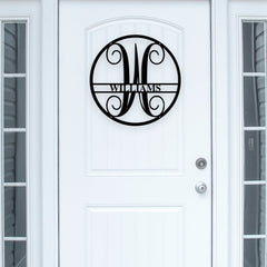 A black metal circular monogram wall art piece featuring an ornate 'M' and the name 'WILMANS' across a horizontal bar, mounted on a white paneled front door.