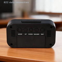A black digital alarm clock shown from the back, resting on a wooden table with a blurred indoor background. White text at the top reads '822 daily necessities'.