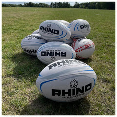 Several white rugby balls with black and blue branding reading 'RHINO AVALANCHE' are stacked on a grassy outdoor field.
