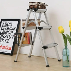 A three-step aluminum step ladder with black rubber feet, shown in a styled room setting with a framed alphabet poster, books, a basket, and yellow tulips in a glass vase.