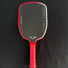 A red and black pickleball paddle with a textured red grip handle, photographed flat against a black fabric background.