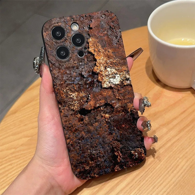 A hand holds a smartphone case with a highly textured, distressed rusted metal appearance in dark brown and orange tones, set against a background of a wooden table and a white coffee cup.