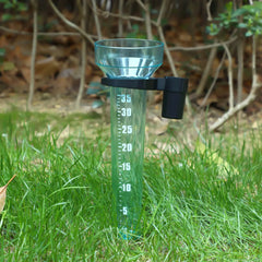 A transparent plastic rain gauge with a black spout and white measurement markings, placed in green grass outdoors with trees in the background.