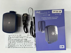 A wearable wireless speaker set with its packaging, manual, and charging cable on a white surface. The product is dark blue with a metallic mesh grille, shown next to its purple retail box and instruction sheet.