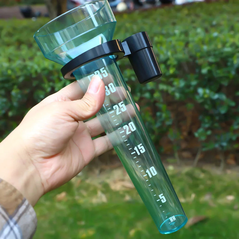 A hand holding a clear green plastic rain gauge with white measurement markings and a black cap, set against a blurred green garden background.