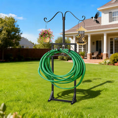 A black metal garden hose stand on a green lawn in front of a house, holding a coiled green garden hose. The stand has curved hooks with hanging planters and bird feeders, and a bird is perched on one of the feeders.