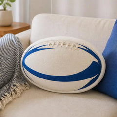A white rugby ball-shaped throw pillow with blue curved stripes and faux laces, resting on a beige sofa next to a blue pillow and a gray throw blanket.