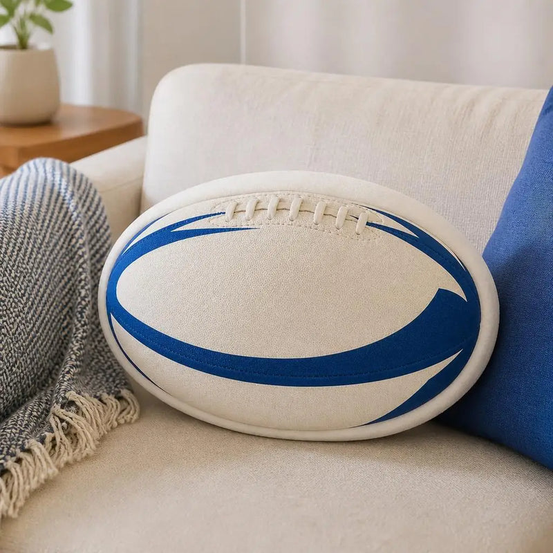 A white rugby ball-shaped throw pillow with blue curved stripes and faux laces, resting on a beige sofa next to a blue pillow and a gray throw blanket.
