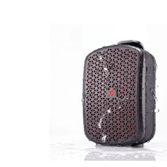 A rugged black portable speaker covered in water droplets, standing on a reflective white surface against a white background.