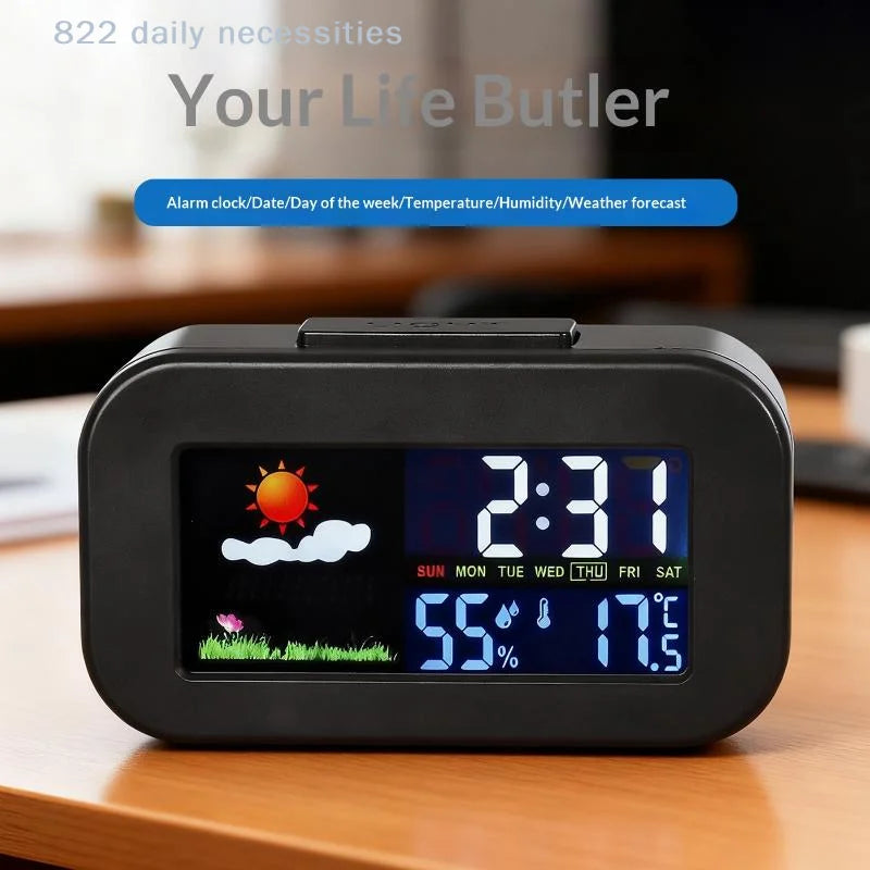 A black digital alarm clock with a colorful LCD display sitting on a wooden table. The screen shows the time 2:31, weather icons, temperature 55 degrees Fahrenheit, and humidity 70%.