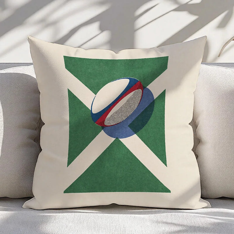 A square throw pillow with an off-white background featuring a green saltire design and a stylized rugby ball graphic in the center, resting on a light-colored sofa with dappled sunlight.