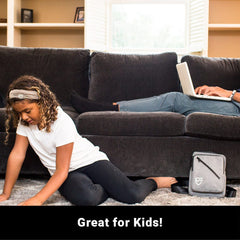 Child playing with a gray bag on the floor, with an adult using a laptop on a couch in the background.